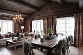 Grey fur blankets on chairs around solid wooden table and comfortable lounge area in open-plan interior of rustic wooden house