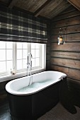 Freistehende Badewanne vor Fenster, oberhalb Raffrollo mit Karomuster, in Bad eines Holzhauses