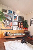 Dog on flokati rug and couch with ethnic blanket against half-height, continuous sideboard below collection of pictures on wall