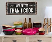 Colourful bamboo bowls and container with lid on wooden lowboy below sign with humorous motto on wall