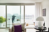 White table lamp with domed lampshade on side table next to armchair with scatter cushion; glass wall with panoramic view