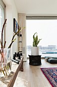 Branches in glass vase on rustic wooden bench, house plant in white planter on lattice table and glass wall with view of harbour entrance