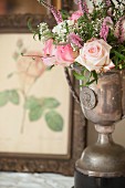 Rosenstrauss in antiker silberner Pokalvase, im Hintergrund Bilderrahmen mit Blumenmotiv
