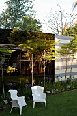 Two white, postmodern plastic chairs in front of mirrored façade and leopard trees in herbaceous border