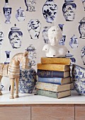Bust of woman on stacked books, blue and white painted ceramic vase and bowls in front of wallpaper with pattern of vases