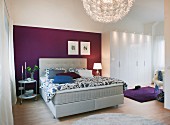 A bedroom with a light grey boxspring bed against a purple wall with a white, multi-door wardrobe to the side
