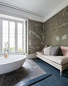 Oval, free-standing bathtub on stone tiles below window and antique couch against patinated wall