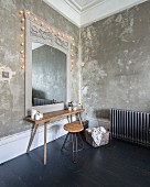 Rustic wooden table and retro stool below mirror with antique frame on patinated wall and decorated with fairy lights