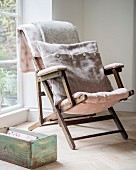 Vintage-Gartenstuhl mit altrosa Polsterung und Wollstoff-Kissen vor Fenster, davor alter Holzkiste auf Eichenparkett