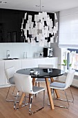 Dining table with black, round top and white chairs below Zettel'z lamp in open-plan kitchen