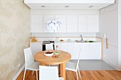 Runder Holztisch und weiße Stühle vor Küchenzeile mit Hängeschränken in Weiß