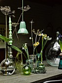 Various flowers in vases and turned candle in candlestick next to vintage, green pendant lamp