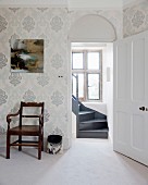 Antique armchair against wall with ornate wallpaper and view of winding wooden staircase through open door
