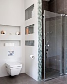 Wall-hung toilet below shelves in niches next to shower cubicle with glass door