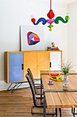 Postmodern pendant lamp with colourful arms above dining table with wooden top mounted on metal frame, metal armchairs and sideboard with colourful doors in background