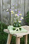 Delicate campanula and ox-eye daisies in green glass bottles on folded green and white tablecloth on rustic wooden stool