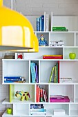Books and colourful ornaments on white shelves; yellow pendant lamp in foreground