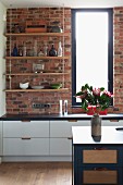Open-fronted, suspended shelves against brick wall above white base units and blue island counter in open-plan kitchen