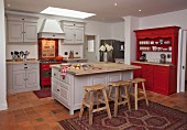 Wooden bar stools at free-standing island counter in open-plan, country-house kitchen in shades of pale grey and red with recessed spotlights in suspended ceiling