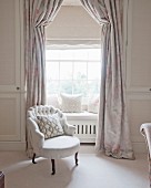 White armchair in front of bay window with window seat and elegant curtains