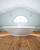 Moderne, freistehende Badewanne auf rustikalem Dielenboden, vor Giebelwand mit halbrundem Fenster