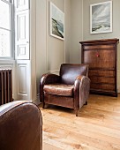 Brown, vintage leather armchair next to window in front of narrow chest of drawers against wall painted pale grey in traditional interior