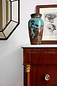 Painted, antique vase on cabinet in front of framed drawing leaning against wall