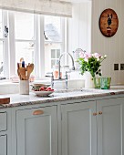 Küchenzeile vor Fenster, Arbeitsplatte aus Stein auf hellgrau lackierten Unterschränken im Landhausstil