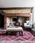 Ottoman and antique armchair in front of log-burner in old, stone inglenook fireplace