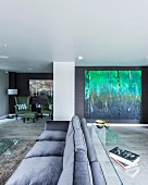 Grey three-seater sofa with narrow glass table against backrest in spacious living room with large painting of forest on wall
