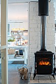 Fire in vintage-style log-burner against white brick wall and view into kitchen