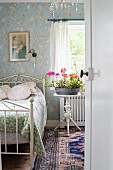 Blick durch offene Tür ins Schlafzimmer mit romantischem Flair, auf Beistelltisch Topf mit blühenden Blumen neben Bett mit verziertem Metallgestell in Weiß
