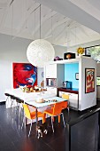 Designer pendant lamp above dining table and orange chairs next to free-standing counter and bar stools in open-plan kitchen
