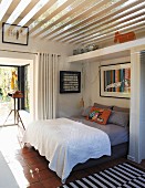 weiße Tagesdecke auf Doppelbett in offenem Schlafbereich, oberhalb Glasdach mit Holzlamellen Abdeckung