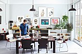 Man in dining area with dark wooden chairs around modern table and collection of pictures on wall above open-fronted sideboard in loft apartment