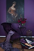Armchair with purple covers and vase of red flowers on table below oil painting on aubergine wall