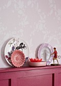 Decorative plates and figurines on a wooden shelf against a wall with pink and white vine patterned wallpaper