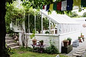 Gewächshaus mit Holzverschalung in Weiß, davor Blumentöpfe auf Tisch und Truhe in sommerlichem Garten
