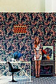 Young woman in room with different patterns on wallpaper, carpet, lampshade and dress