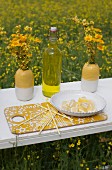 Slices of lemon on white plate and swing-top bottle between vases of dyer's chamomile on white, vintage surface in field of flowering rapeseed
