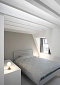Weißes Bett mit eleganter Tagesdecke in Dachschlafzimmer mit sichtbarer weißer Balkenkonstruktion und Gaubenfenster