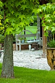 Gartenbank auf Natursteinbelag, im Vordergrund Ahornbaum, in sommerlichem Garten