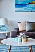 Round retro coffee table in front of sofa with scatter cushions and table lamp on side table below painting on wall