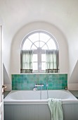 Badezimmer mit Badewanne vor teilweise gefliester Wand und Rundbogenfenster