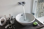 A wash stand with a bowl basin with water flowing into it from a wall mounted designer tap