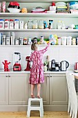 Girl on step stool in front of kitchen counter and open shelf full of storage containers