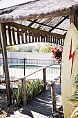 Wooden jetty with thatched roof and surfboard ajar, pool view