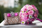 Romantic, pink bridal bouquet next to elegant handbag with sparkling brooch on top of vintage suitcase