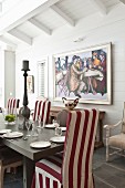 Set dining table and red and white striped loose-covered chairs in front of framed painting
