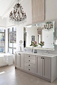 Pale grey bathroom washstand below elegant mirror, chandelier and high, white-painted roof structure